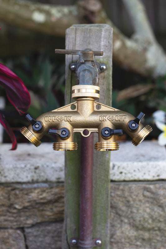 Brass Four Way Tap Adapter attached to an outdoor garden tap, mounted on a wooded post in front of a garden bed