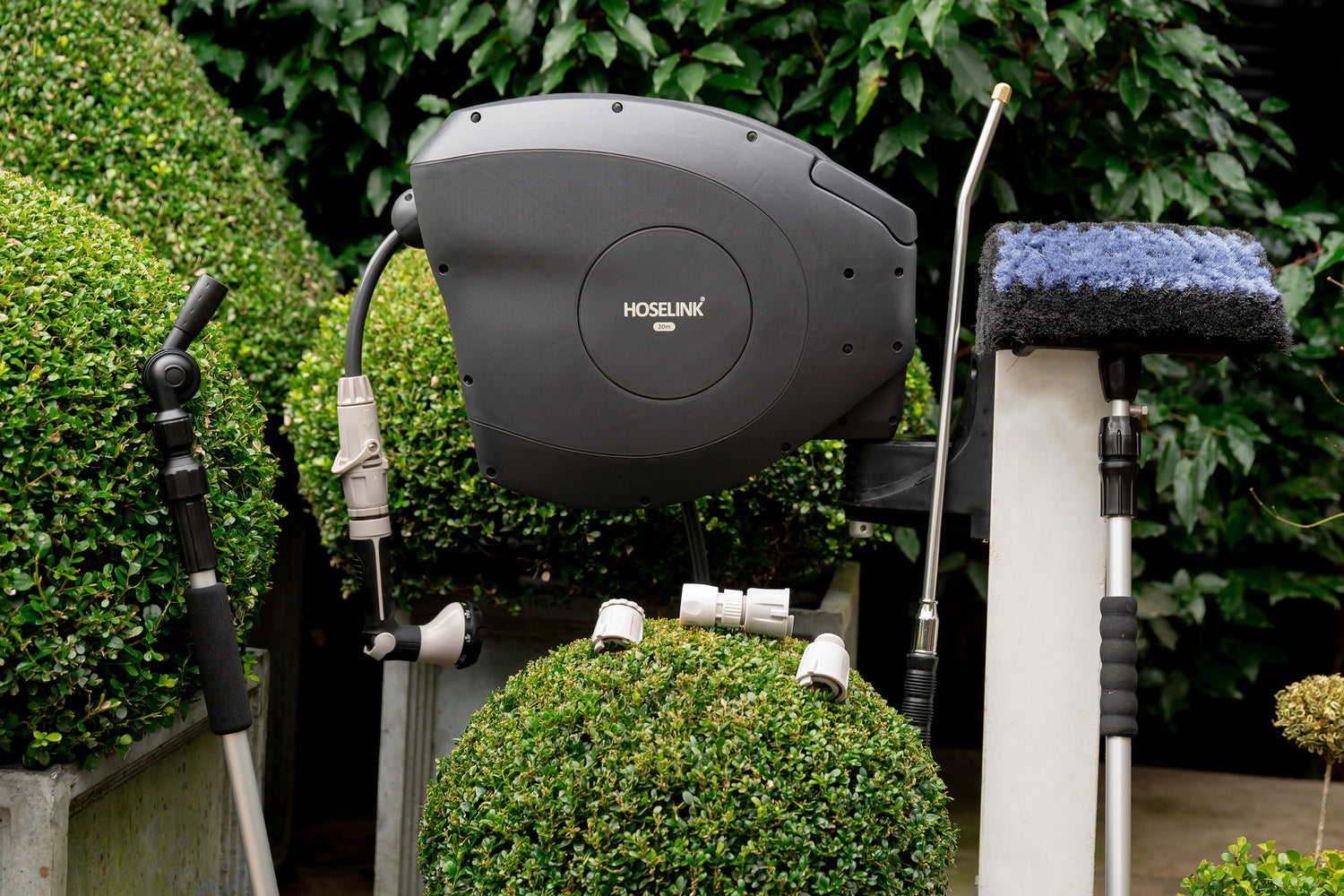 Charcoal Retractable Hose Reel mounted to a white timber post in a garden surrounded by the Hoselink cleaning brush, connectors, the pivot gutter cleaner and super jet washer 