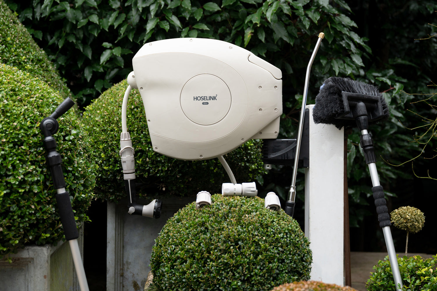 Beige Retractable Hose Reel mounted to white timber post in garden surrounded by cleaning brush, connectors, the pivot gutter cleaner and super jet washer 