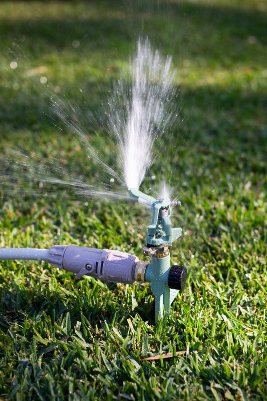 A close up image of a pale green Impulse Sprinkler spiked into a lawn and connected to a hose with Hoselink connectors, in the middle of spraying water from the impact spray head.