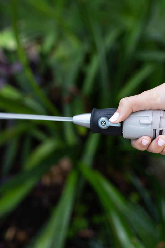 A close up image of a woman's hand holding hose connectors and a short black nozzle with a lever, spraying a jet stream of water in front a a dark green garden background.