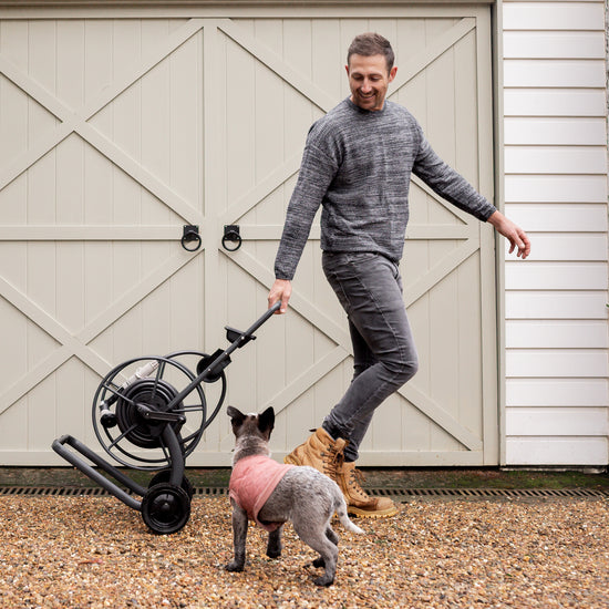 man using charcoal metal hose reel cart as dog follows