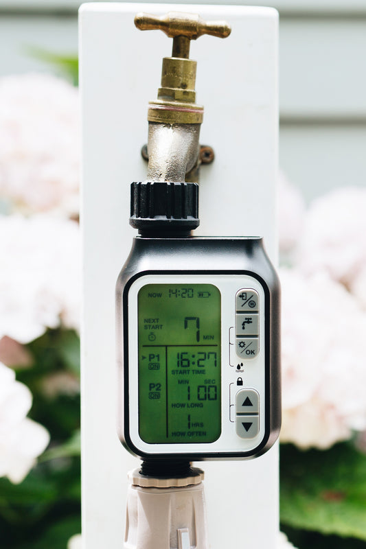 A timer with a digital LCD display and five buttons connected to a garden tap with hose connector underneath and pale pink flowers in the background.