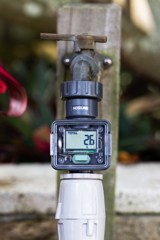 A water flow meter gauge with small digital display and one button connected to a tap with hose connectors joined underneath.