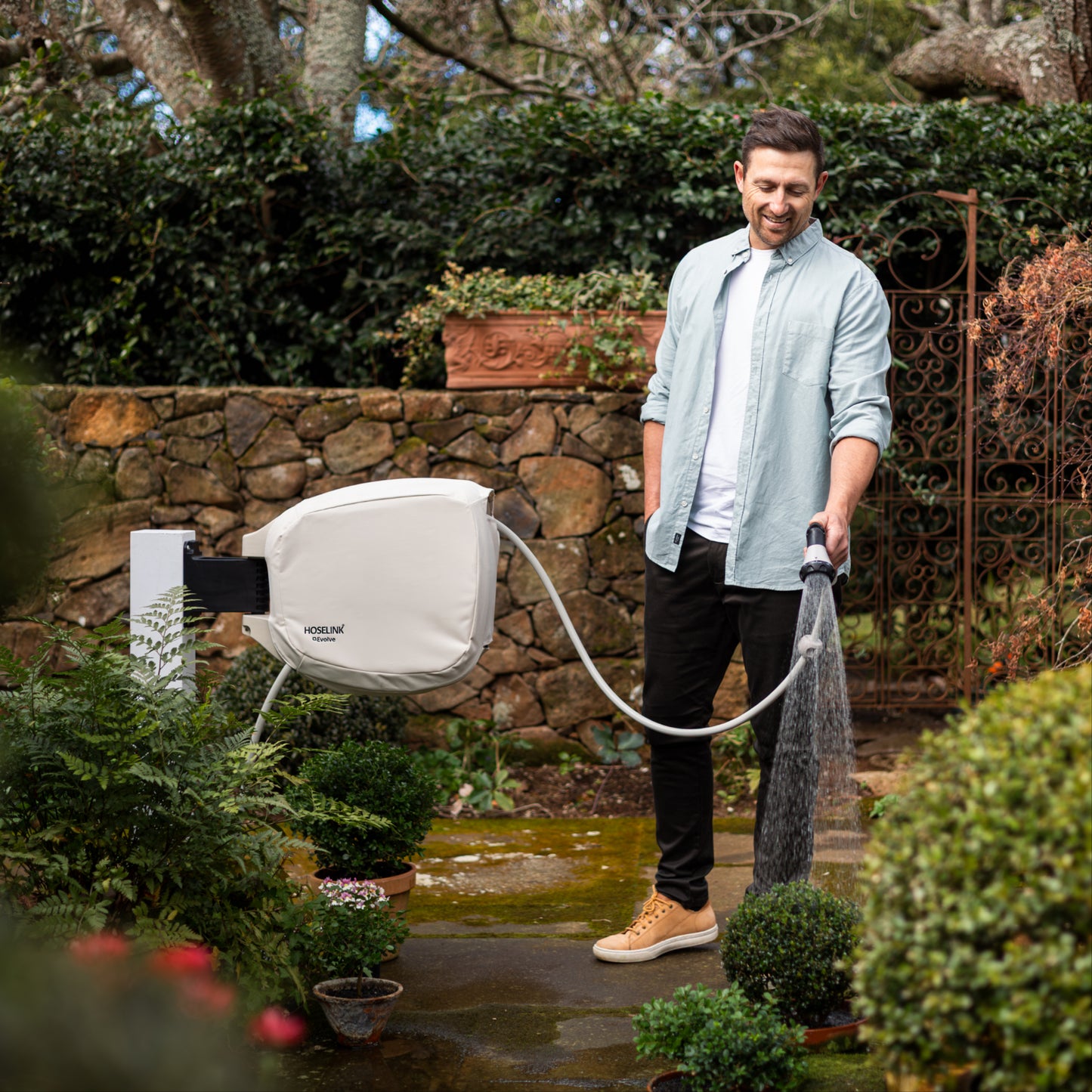 Man using evovle hose reel in beige cover to water plants in a garden with a stone wall and trees in the background.
