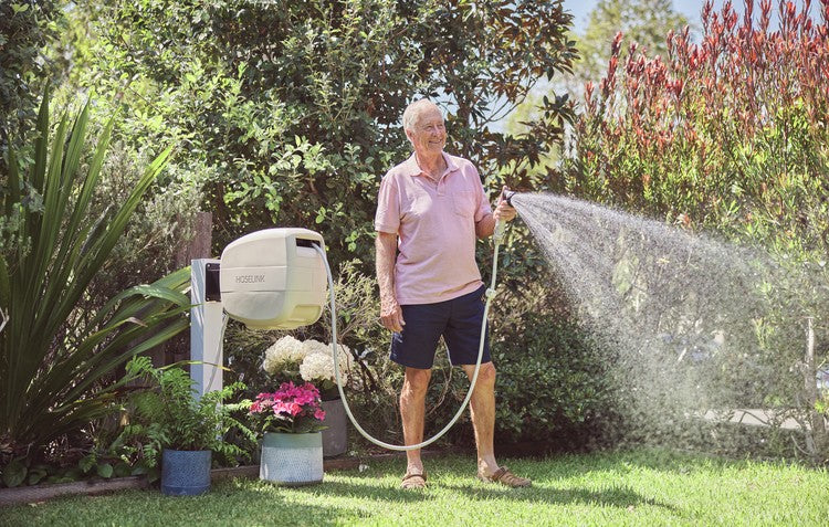 older man wearing a pink shirt watering his garden with a retractable hose reel