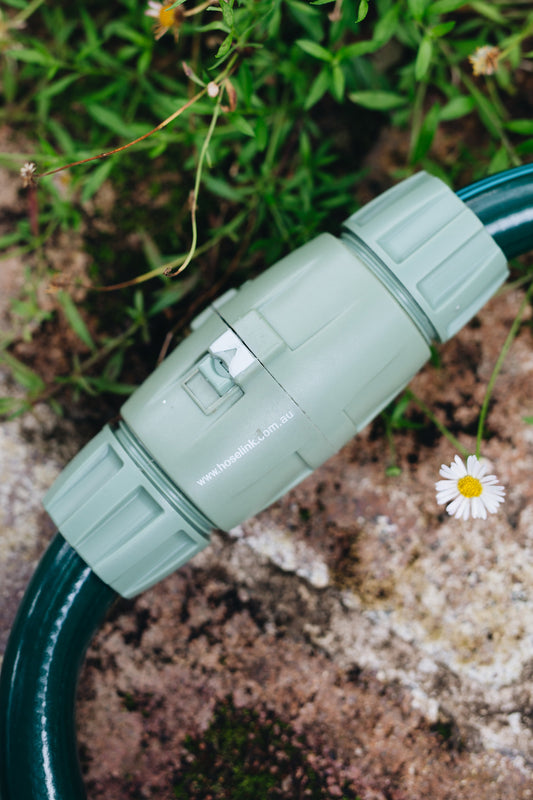 Two light green 18 millimetre Hose End Connectors joining a dark green hose laying across sandstone bricks with white daisies around.