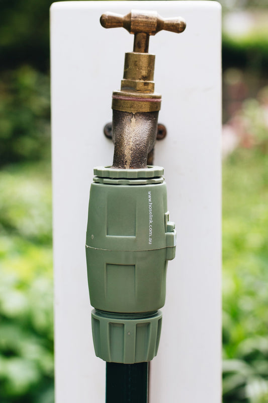 Light green 18 millimetre Universal Tap Connector and Hose End Connector, connecting dark green hose to a brass outdoor tap which is mounted on a white wooden post in a garden.