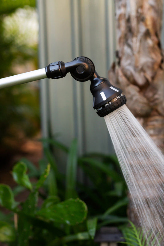 A close up image of the head of a spray wand titled at an angle with a shower spray being emitted, in a garden.