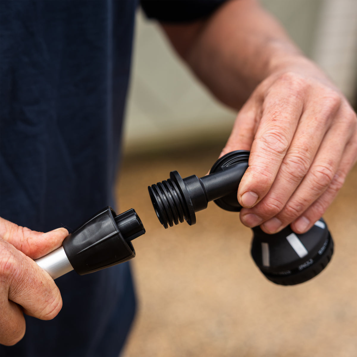 Close-up of a person's hands unscrewing a black plastic garden hose nozzle attachment from a hose, with a blurred outdoor background.