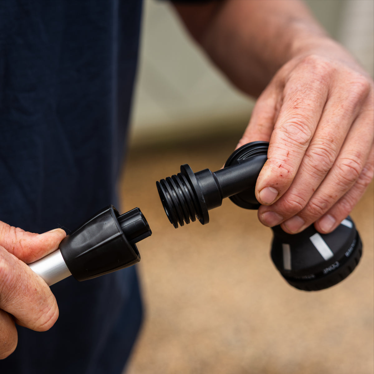 A close-up of a person's hands holding  black plastic 8-pattern sprayer head attachment, being installed on the silver metal telescopic claning system poler, against a blurred outdoor background.