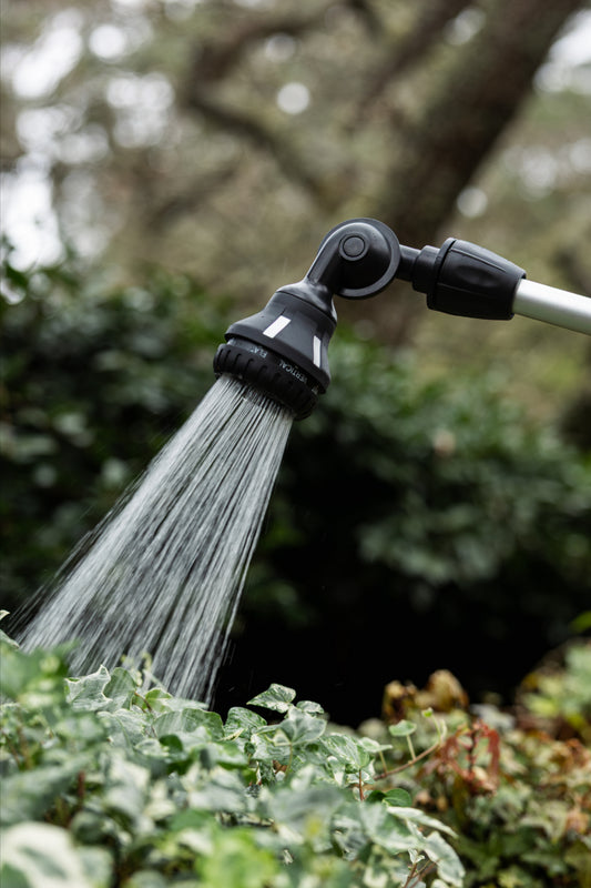 A black adjustable garden hose sprayer with a flexible head spraying water, attached to a silver pole in an outdoor garden setting with blurred green foliage.