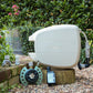 Beige evolve hose reel, 9-pattern sprinkler, automatc tap timer, easy turn 2-way adapter and click-on adapter set sitting on a low brick garden wall surrounded by gravel with a green hedge in the background