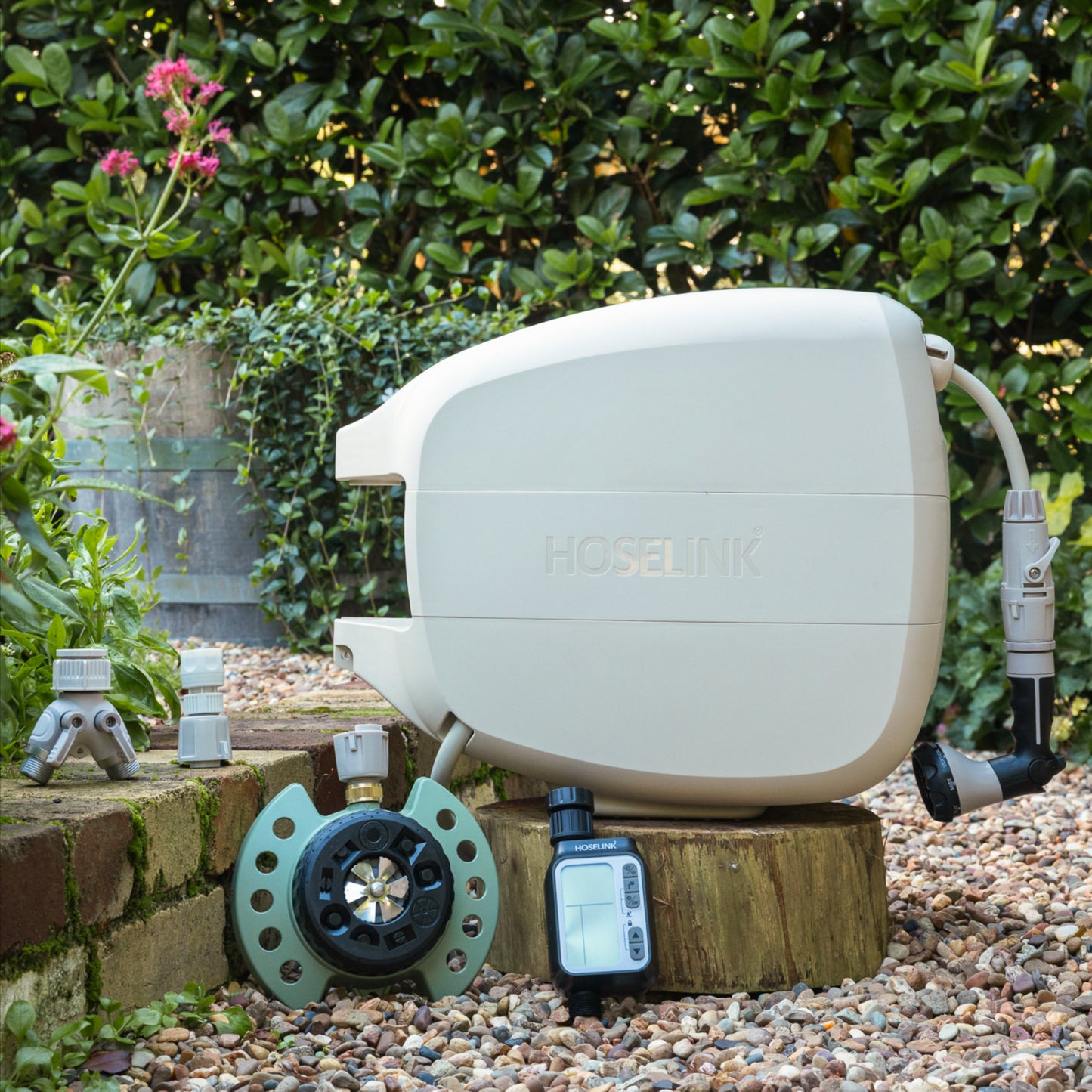 Beige evolve hose reel, 9-pattern sprinkler, automatc tap timer, easy turn 2-way adapter and click-on adapter set sitting on a low brick garden wall surrounded by gravel with a green hedge in the background