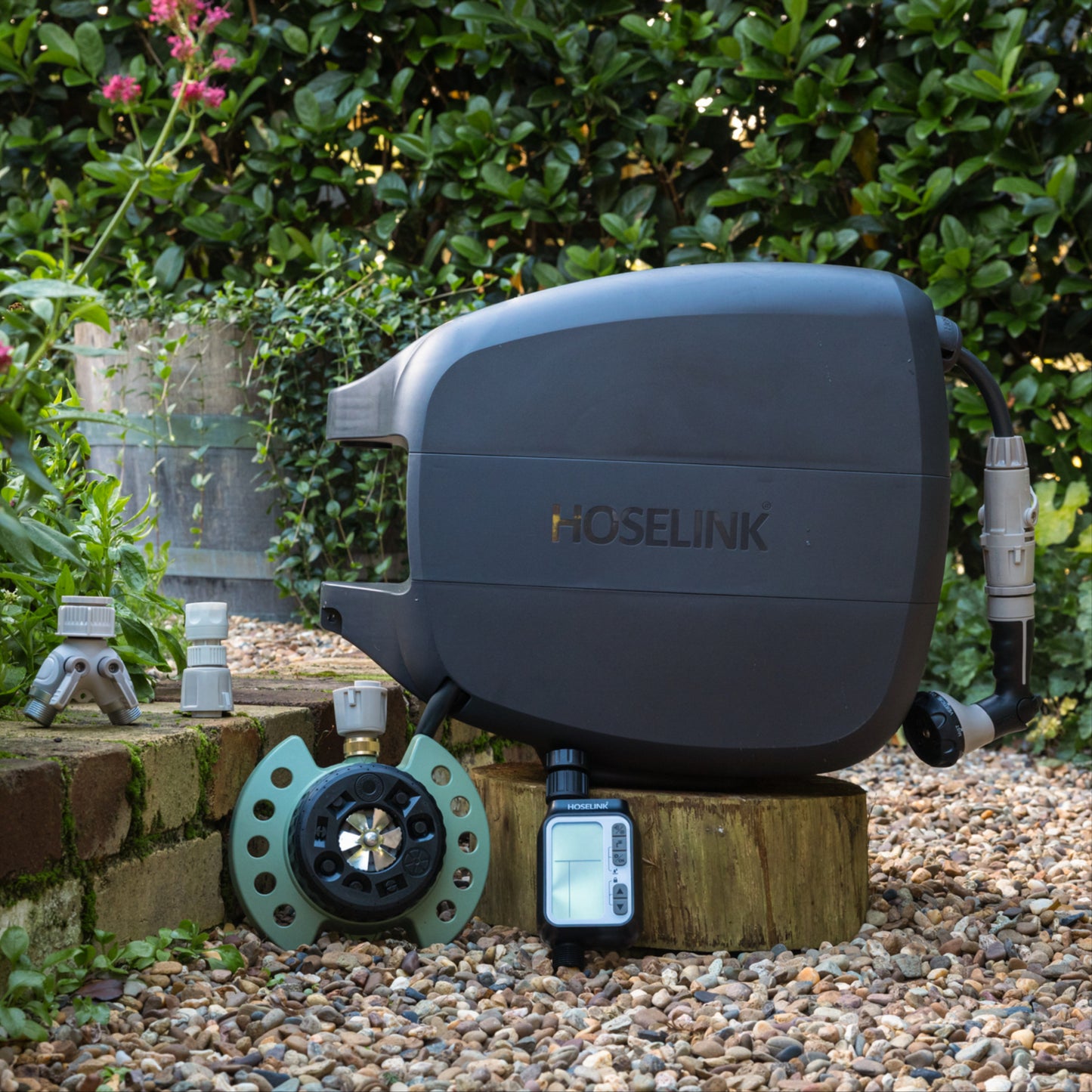 Charcoal evolve hose reel, 9-pattern sprinkler, automatc tap timer, easy turn 2-way adapter and click-on adapter set sitting on a low brick garden wall surrounded by gravel with a green hedge in the background