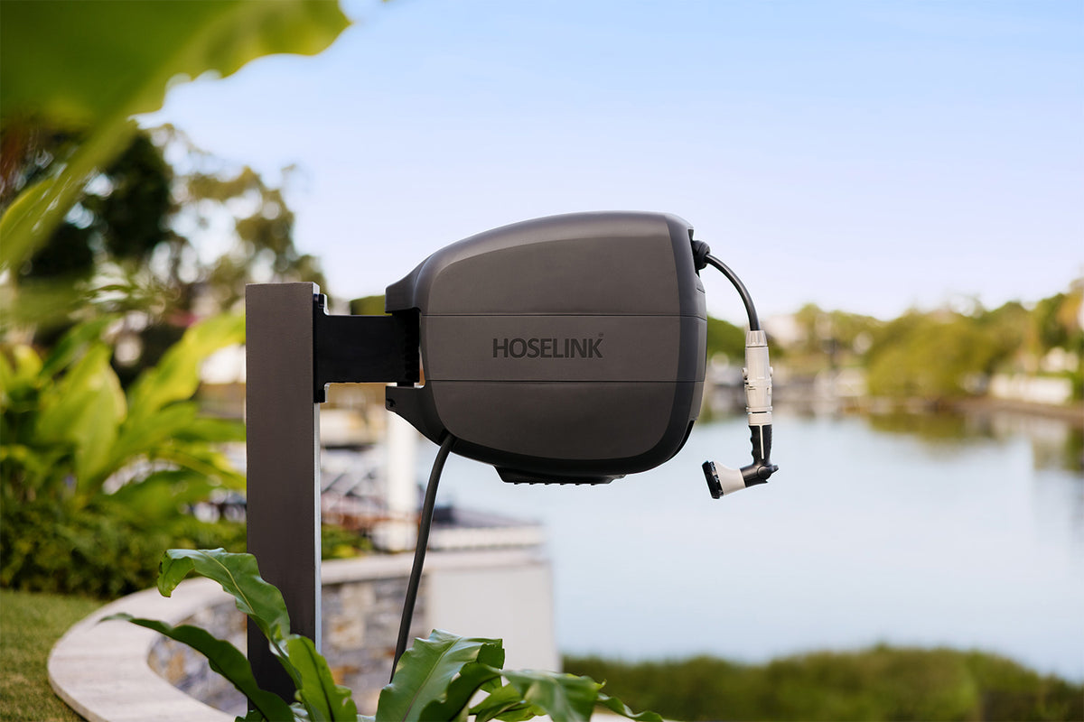 A HOSELINK outdoor reel mounted on a post in a garden setting with a lake or river and trees in the background.