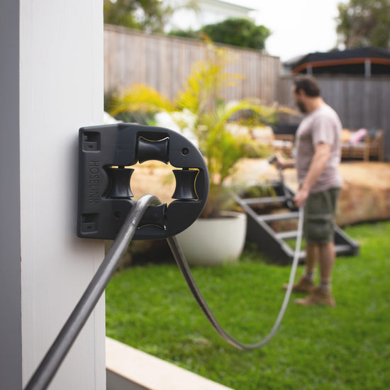 Charcoal-coloured hose being pulled through a Hoselink Hose Guide mounted to the corner of a post while male waters plants in the background