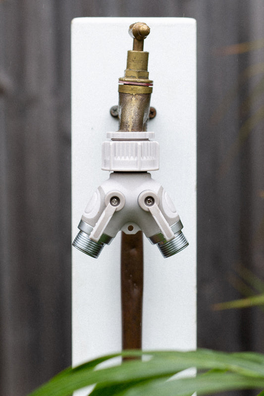 Easy Turn Two Way Tap Adapter on a brass outdoor garden tap, mounted to a white timber post in front of a timber fence