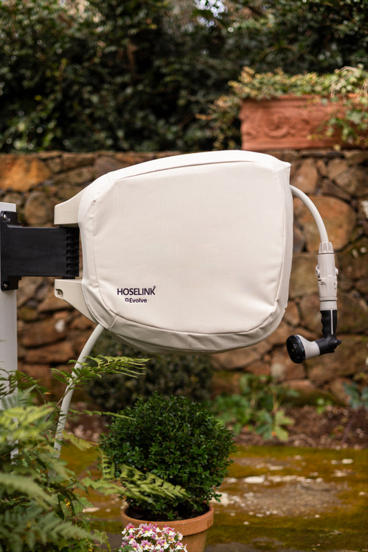 Hoselink Evolve™ hose reel cover on hose reel, mounted outdoors with plants and a stone wall in the background.