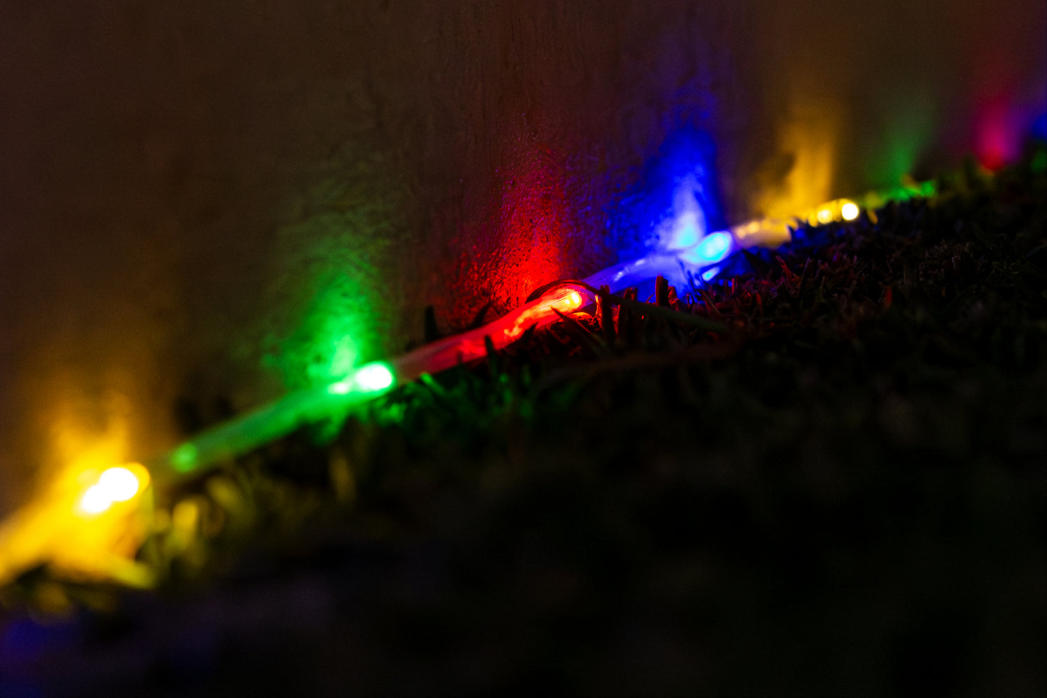 Multicolor LED rope lights glowing on a dark grassy surface against a textured wall, showing red, green, blue, green and yellow light reflections.
