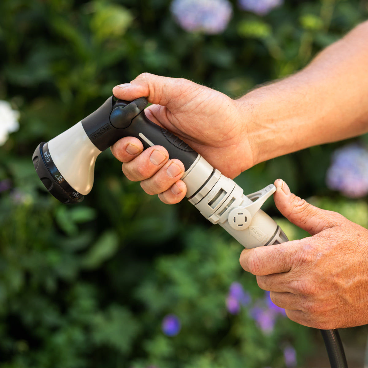 A close-up of a person's hands holding a black and grey garden hose nozzle attachment, with a blurred garden background of green foliage and purple flowers.