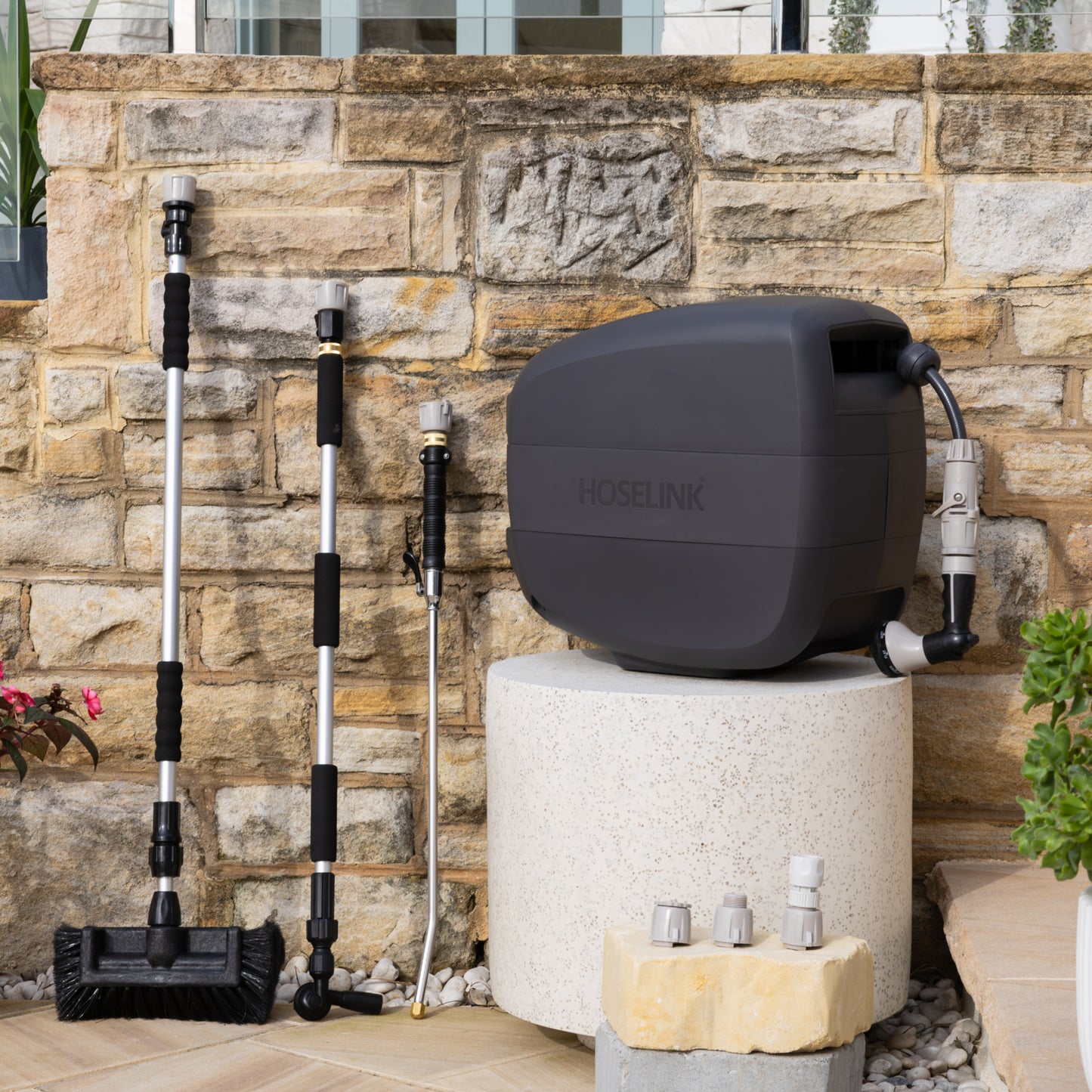 Charcoal Evolve Retractable Hose Reel on a white stone table with an Extendable Cleaning Brush, Super Jet Washer and Pivot Gutter Cleaner leaning on a sandstone wall next to the reel and an adapter pack in front of the reel