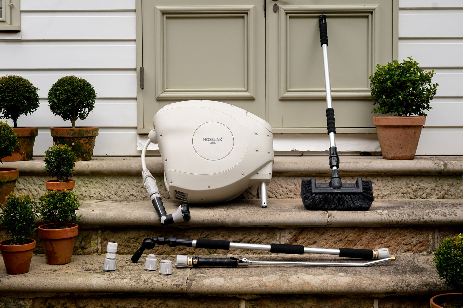 Beige Hose Reel sitting on stone steps outside a house surrounded by Hoselink tools including the Cleaning Brush, Connectors, Pivot Gutter Cleaner and Super Jet Washer
