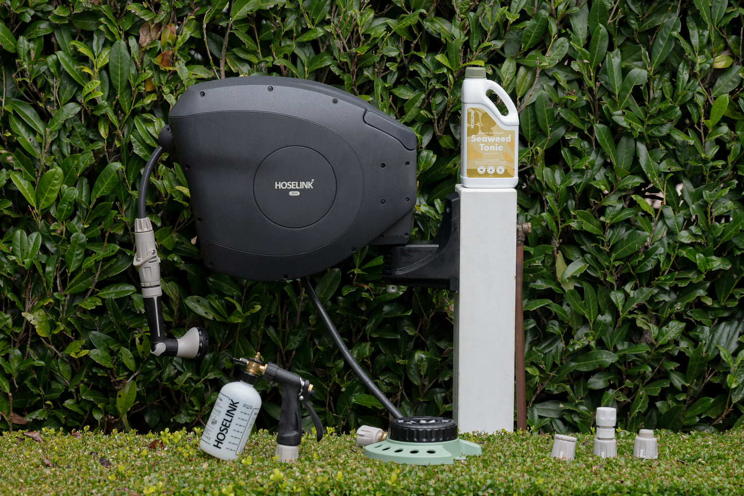 Charcoal Hose Reel mounted to a white timber post in a garden surrounded by Seaweed Tonic, the Fertiliser Spray Mixer, 9-Pattern Sprinkler and Hoselink Connectors