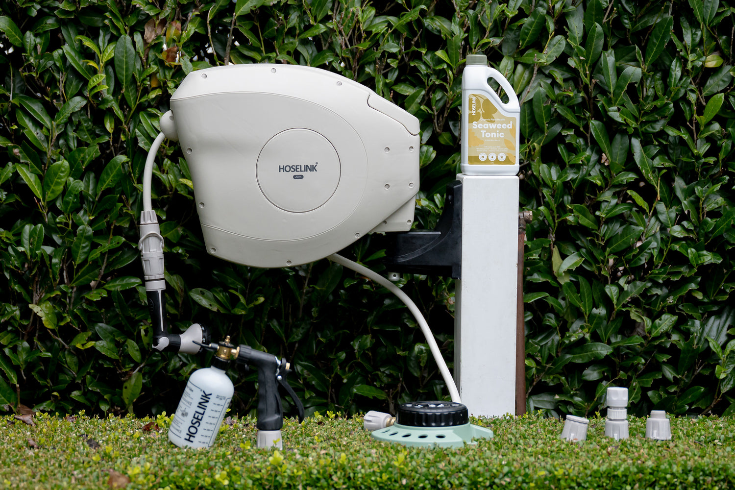Home Gardener Bundle arranged in a garden with Hose Reel mounted on to white timber post and fertiliser, spray mixer, 9-pattern sprinkler and connectors resting on top of the hedge