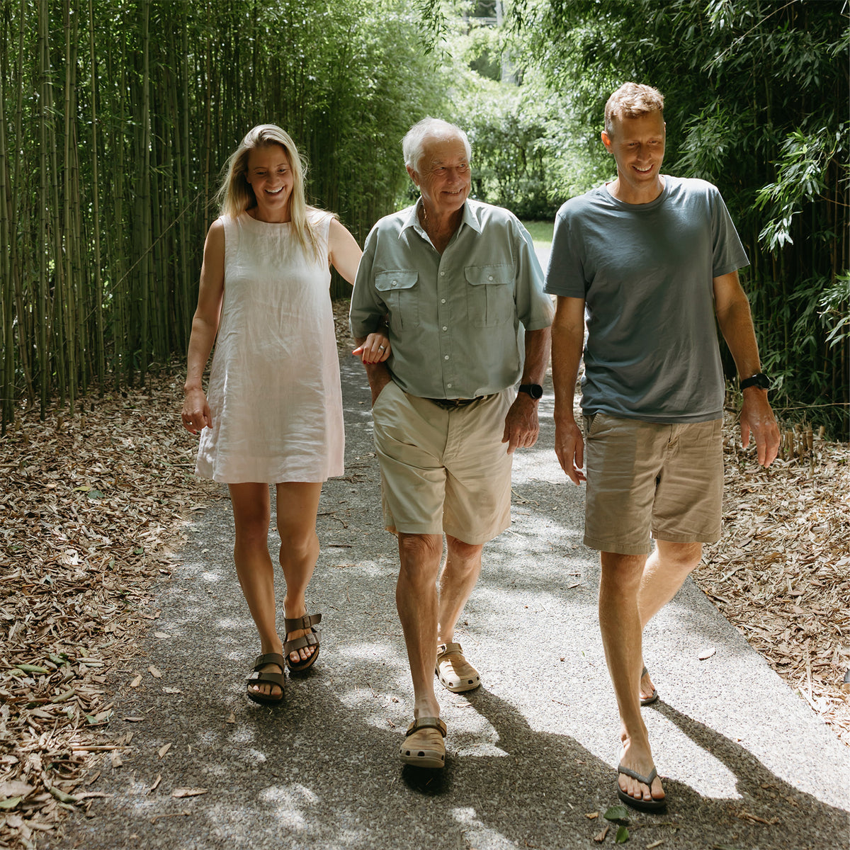 Three people walking arm in arm along a sun-dappled path surrounded by a dense bamboo grove. The woman on the left wears a sleeveless white dress, the man in the center wears a light green button-up shirt and khaki shorts, and the man on the right wears a slate blue t-shirt and khaki shorts.