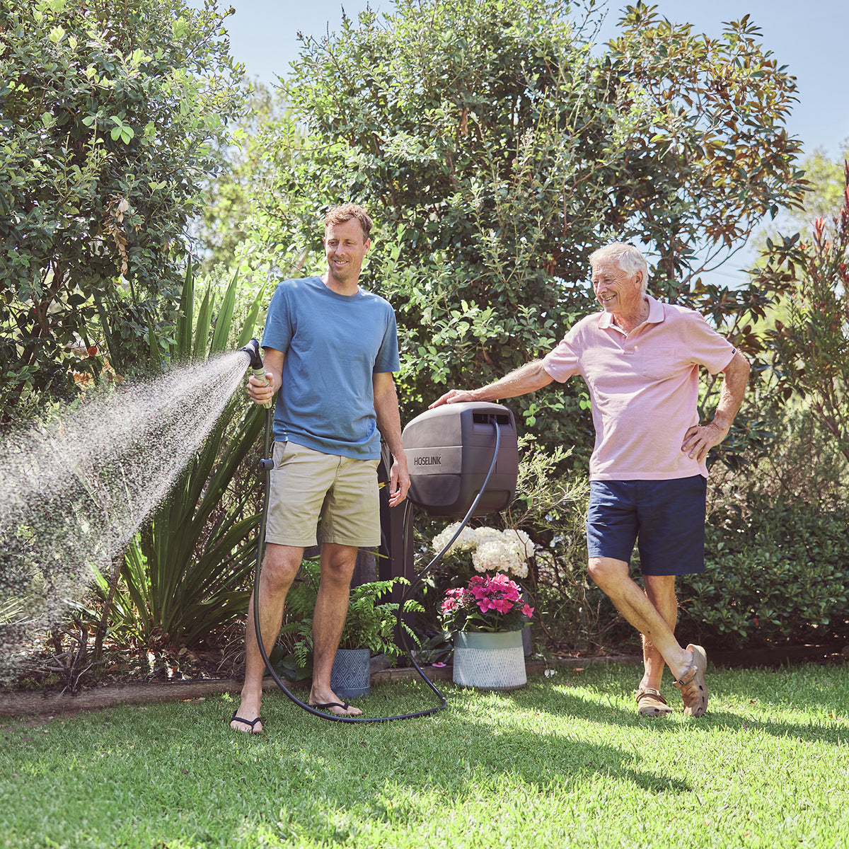A man in a blue t-shirt and khaki shorts waters with a hose while another man in a pink polo shirt and navy shorts watches and smiles in a sunny, green backyard.
