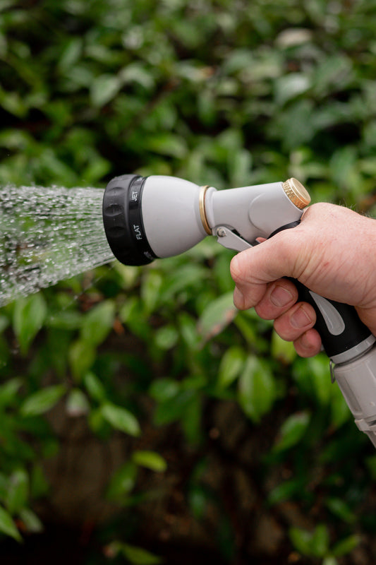 A close up image of a hand holding a black and beige metal multi-function spray gun on a shower setting, with a leafy green background.