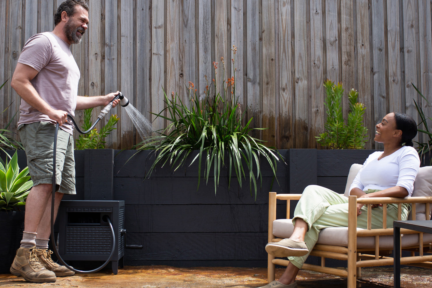 Man Watering His Plants With Charcoal Metal Hose Reel Box With Woman Resting On Outdoor Furniture