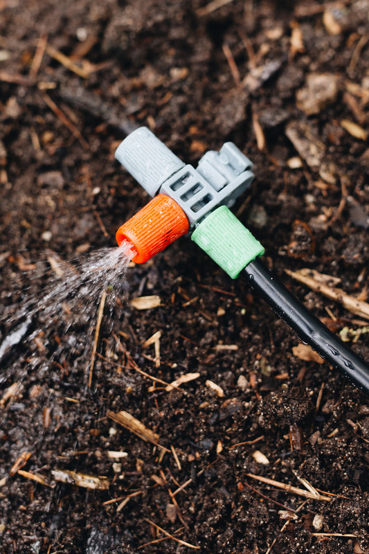 A close up image of a plastic sprinkler head connected to black polypipe over dirt, with a mist spraying from the sprinkler head angled at 180 degrees to the dirt.