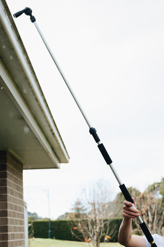 A person holding a long metal pole with rubber grips connected to a hose next to a house, with an angled spray head at the top pointing into the guttering with water spraying.