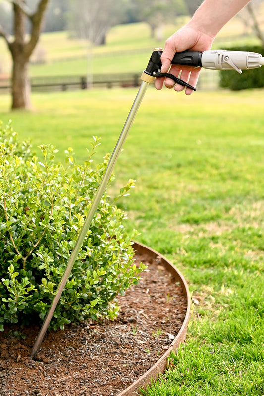 A close up image of a hand holding a trigger spray connected to a hose, with the spray end being a long metal rod pointing into a garden bed at the base of a shrub and lawn in the background.