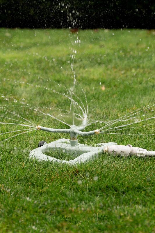 A pale green metal sprinkler on a lawn with a three arm rotating head spraying water in a 360 degree direction.