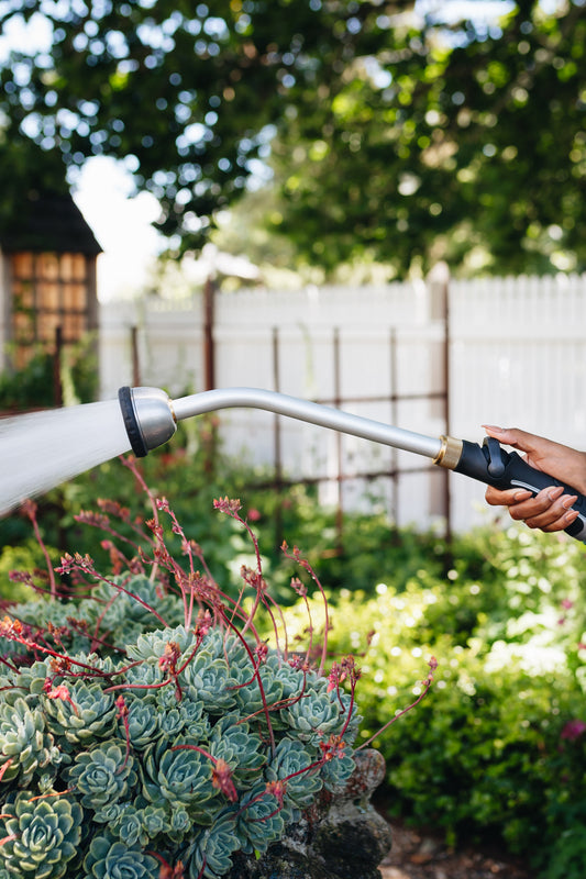 A close up image of a hand holding a long metal spray wand with a lever and water spraying from the shower head, with a fence, small bushes, trees and succulents in the background.