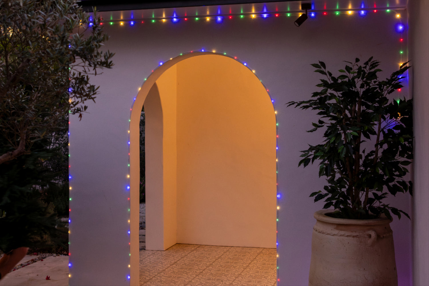 Multicolor LED rope lights draped around a white arched doorway, casting a warm yellow glow, with a potted plant on the right and outdoor foliage on the left.
