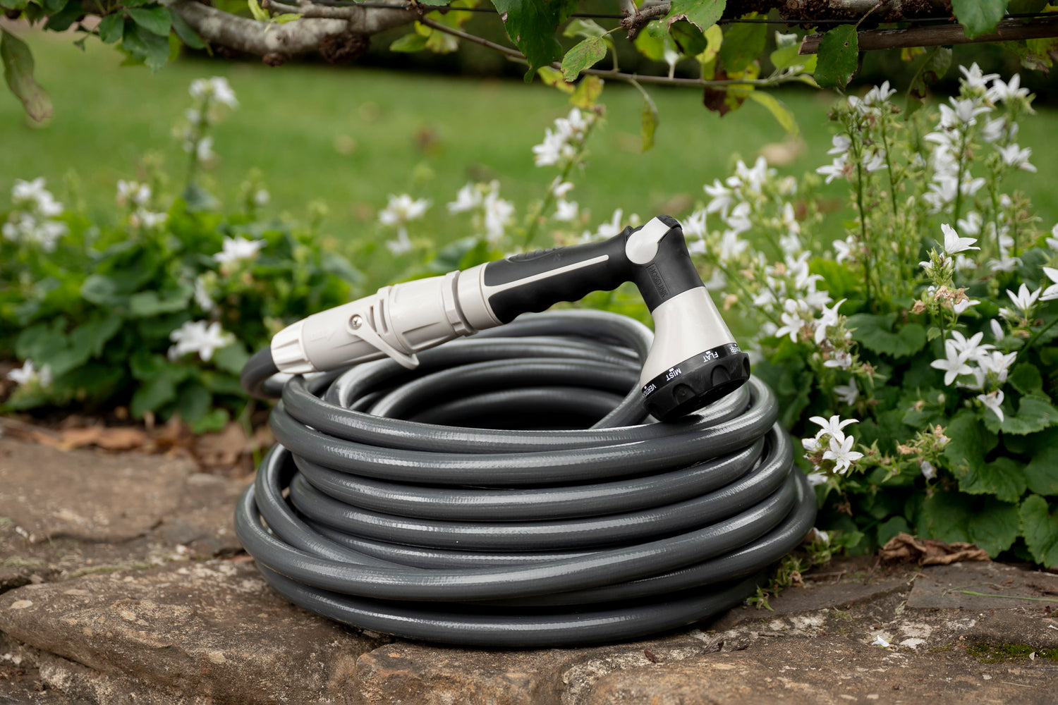 Charcoal-colour Superflex Hose coiled neatly on garden wall with spray gun resting on top, surrounded by blossom.