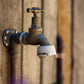 A close-up of an outdoor tap with a grey Hoselink Universal Tap Connector attached to the thread, set against a rustic wooden wall background.