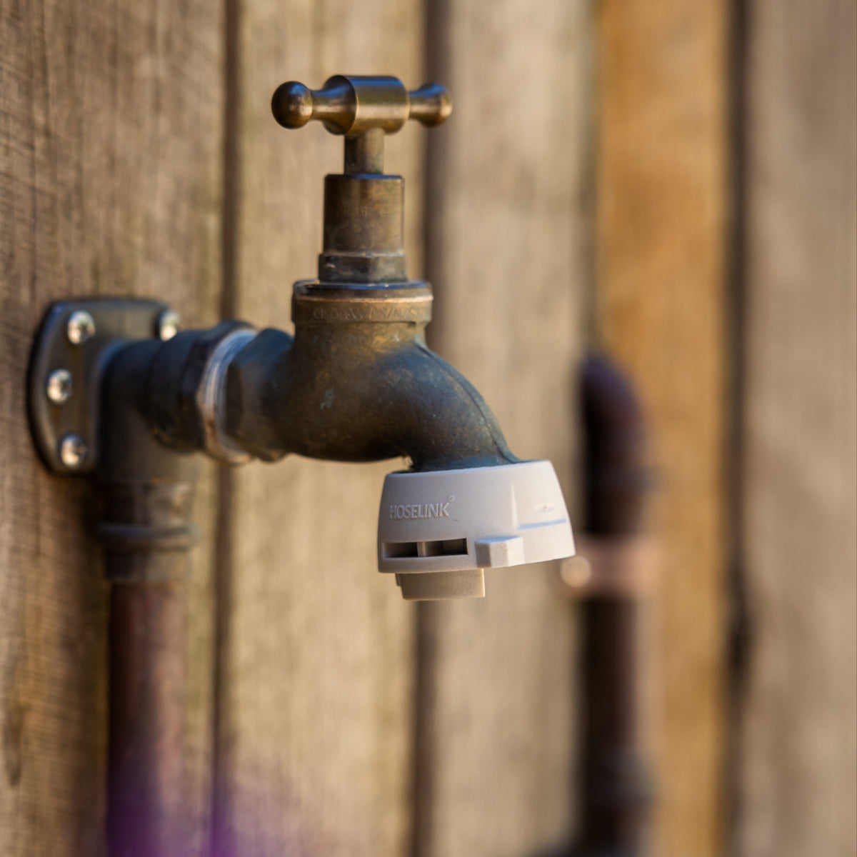 A close-up of an outdoor tap with a grey Hoselink Universal Tap Connector attached to the thread, set against a rustic wooden wall background.