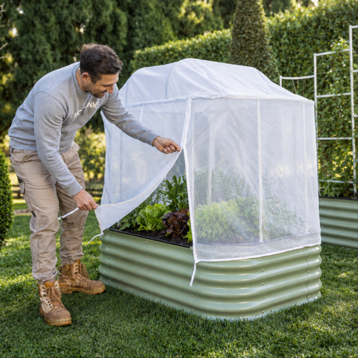 A person opening-the mesh cover attached to a 6 in 1 garden bed in front of a hedge