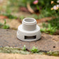 Close up of a grey accessory conneecter with thread sitting on a paved surface with blurred flowers and greenery in the background