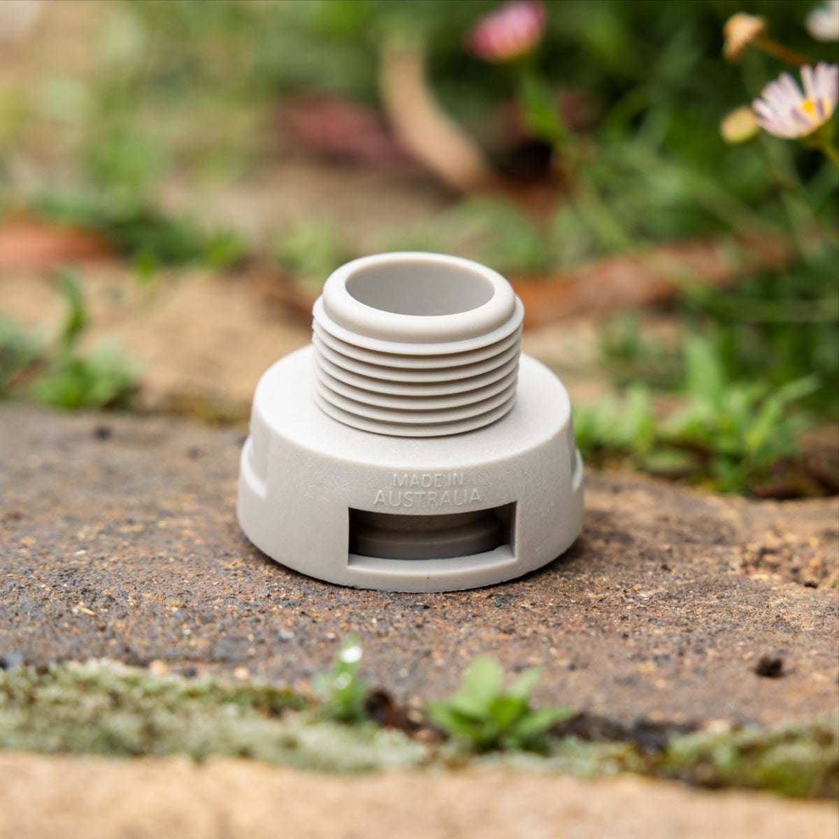 Close up of a grey accessory conneecter with thread sitting on a paved surface with blurred flowers and greenery in the background