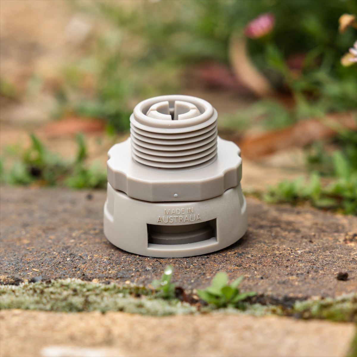 A gray plastic accessory connetor with swivel fitting placed on a stone surface with blurred grass and plants in the background.