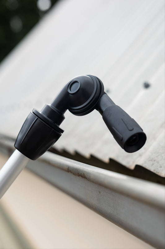 A close-up of a black gutter cleaning head of the telescopic cleaning system attached to a silver pole, infront of a metal roof gutter on a house.
