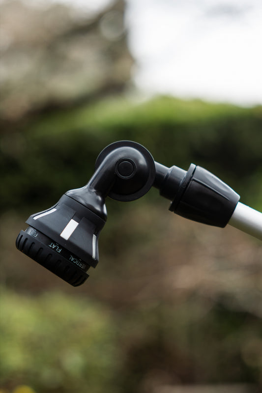 A close-up of a black adjustable garden hose nozzle with a white spray pattern, attached to a silver pole, set against a blurred outdoor background of green foliage.