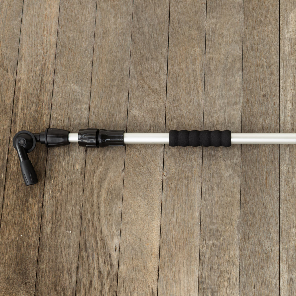 A close-up of a telescoping cleaning system pole with black textured grip sections and gutter cleaner head attachments laid horizontally on a brown wooden plank surface.