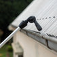 A close-up of a black gutter cleaning head of the telescopic cleaning system attached to a silver pole, spraying water into a metal roof gutter on a house.
